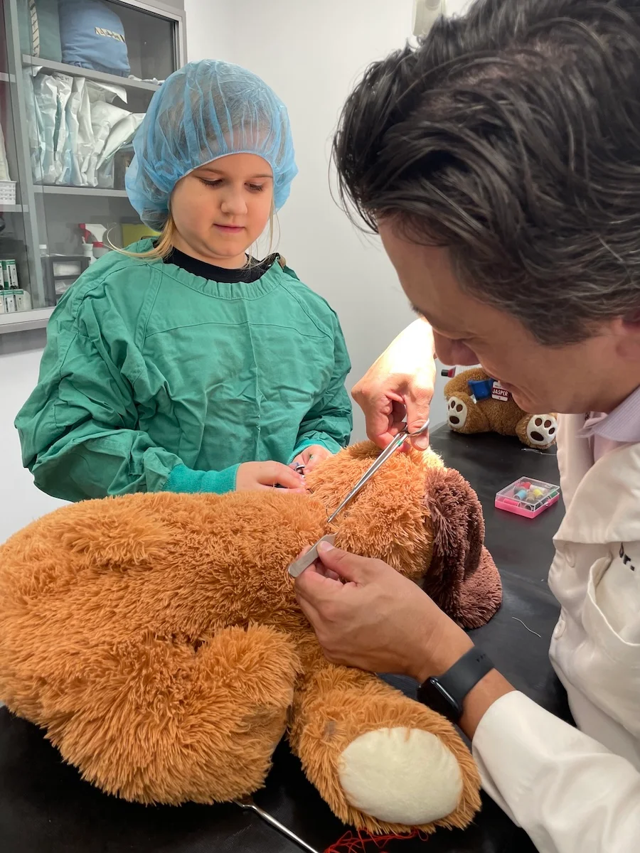 A young girl in surgical attire assisting a veterinarian in suturing a stuffed animal on an exam table.